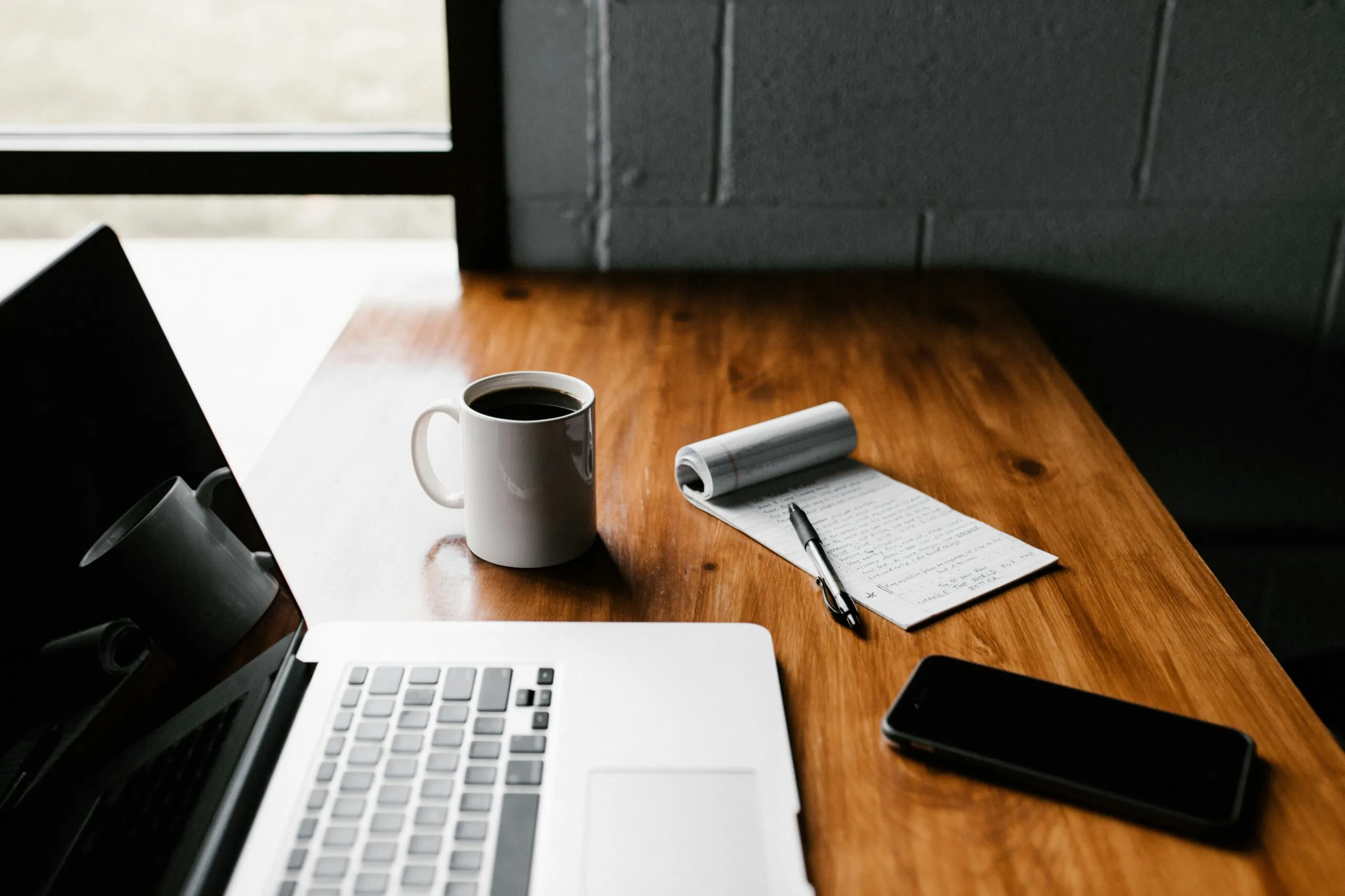 A laptop, a mobile phone, a mug of coffee and a notepad and pen on a desk