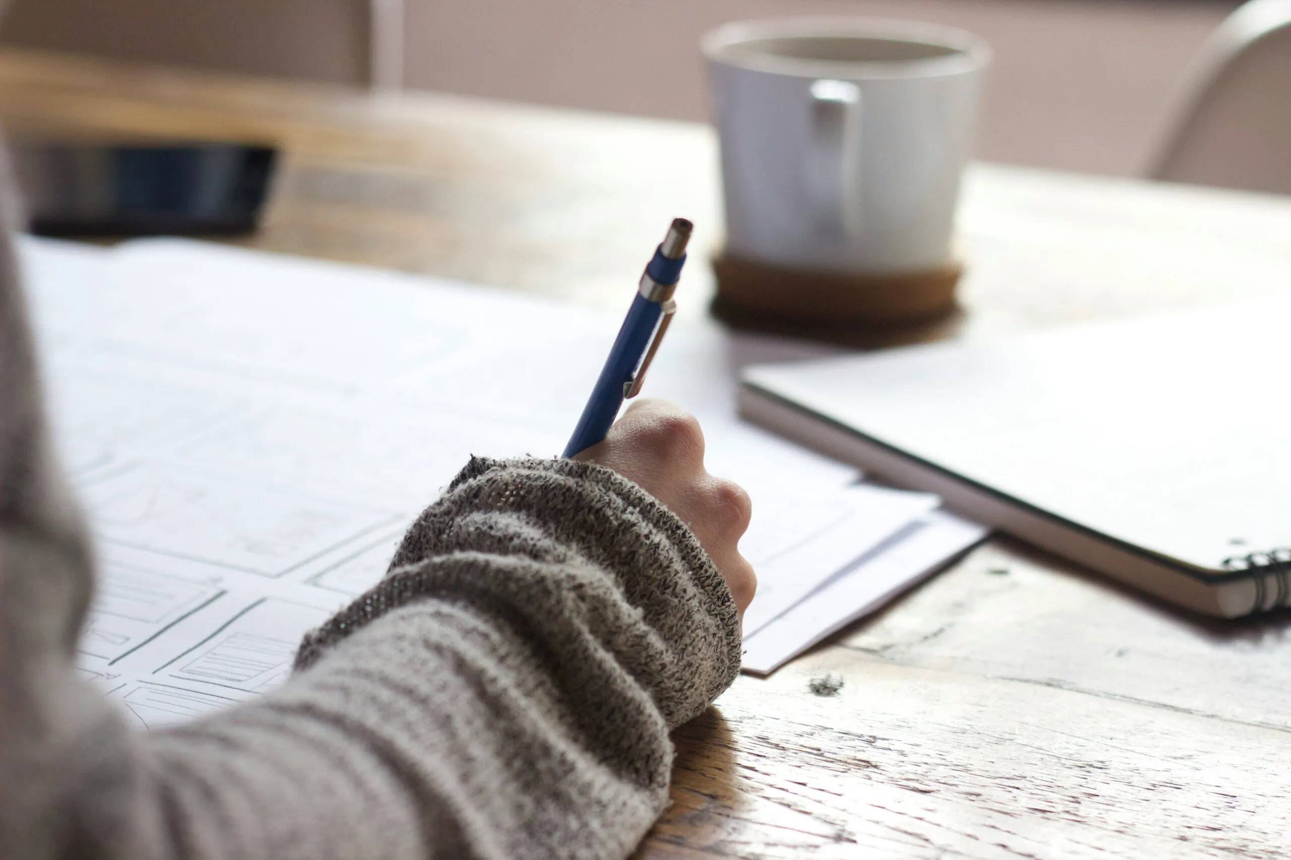 A person writing on paper with a pen, a mug of coffee on the table